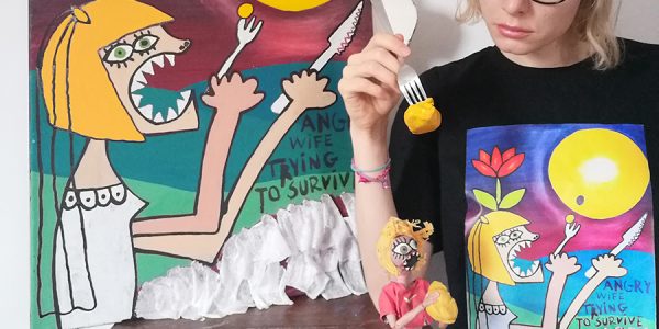 Eine blonde Frau mit einer Gabel in der Hand, auf der eine gelbe Knetmasse aufgespießt ist, vor einem Gemälde, das ihre ungewöhnliche Situation karikierend darstellt. Das gleiche Bild ist zusätzlich auf ihrem T-Shirt abgedruckt und vor ihr als Puppe aufgebaut.