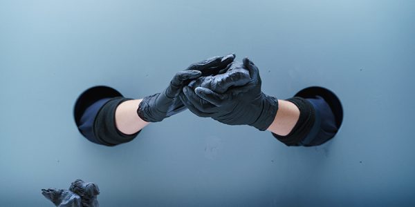 Durch zwei Löcher in einer weißen Wand streckt jemand seine Hände durch und bearbeitet mit schwarzen Handschuhen schwarze Knetmasse.