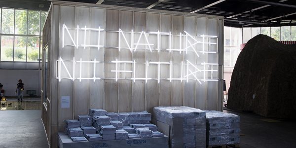 An der Wand eines Holzcontainers stehen in abstrakter Leuchtschrift die Worte „no nature, no future“. Drunter sind Papiere und Platten gestapelt.
