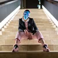 Auf einer breiten geraden Treppe sitzt eine junge Frau mit rosa Schuhen, einem rosa Jumpsuit, einer schwarzen Lederjacke und einer blauen Maske, die das Gedicht versteckt.