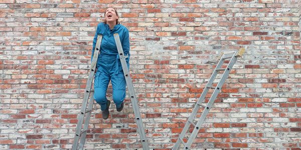 Vor einer alten Backsteinmauer stütz sich eine Person in einem blauen Overall auf zwei Leitern, während die Füße in der Luft baumeln.