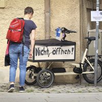 Auf der Kiste eines kleineren Lastenrades steht geschrieben „Bitte nicht Öffnen!“. Eine Frau mit rotem Rucksack steht vor dem Fahrrad und schaut sich die weiteren Hinweise an.