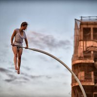 Eine Frau in kurzem weißen Outfit stützt sich auf einem Metallbogen in der Luft. Im Hintergrund sind die Baracke eines Hochhauses und der Himmel mit einzelnen Wolken zu sehen. Sie scheint in der Luft zu schweben