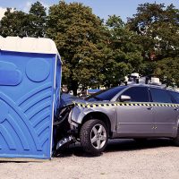 Ein Auto, das frontal durch den Aufprall an einem Dixi-Klo, komplett zerknautscht ist, steht auf einem von Bäumen umgebenen Platz