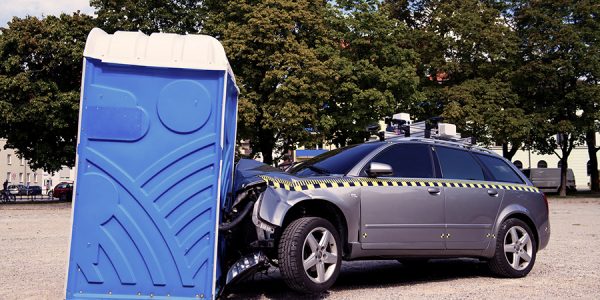 Ein Auto, das frontal durch den Aufprall an einem Dixi-Klo, komplett zerknautscht ist, steht auf einem von Bäumen umgebenen Platz