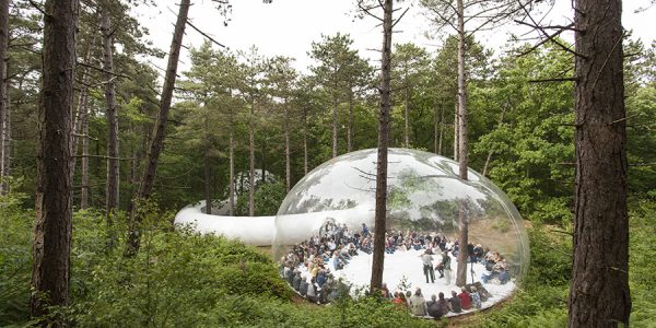 Mitten auf einer Waldlichtung stehen zwei große Luftgefüllte Blasen, um mehrere Bäume herum oder sie einbeziehend. In der größeren Blase sitzen viele Personen und lauschen drei Musikern.