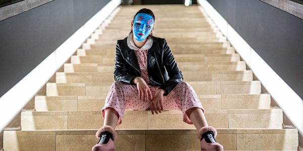 Auf einer breiten geraden Treppe sitzt eine junge Frau mit rosa Schuhen, einem rosa Jumpsuit, einer schwarzen Lederjacke und einer blauen Maske, die das Gedicht versteckt.