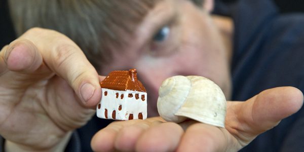 Eine Person hält auf ihrer flach ausgestreckten Hand das Häuschen einer Weinbergschnecke und ein kleines Wohnhäuschen aus Ton.