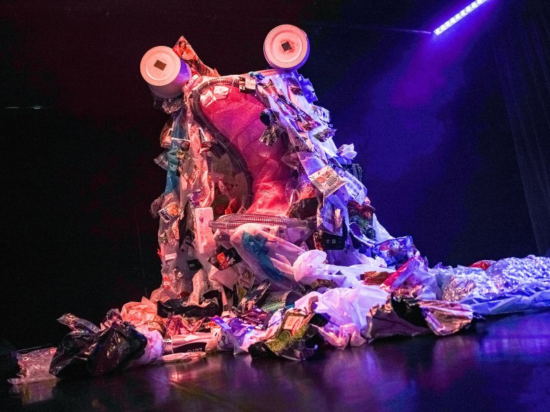 Große Figur, die aus Müll und Plastikabfällen besteht, auf einer Bühne mit lilanem Licht. Die Form erinnert an ein Ungeheuer mit großen, runden Augen und offenem Maul, umgeben von weiteren Abfällen.