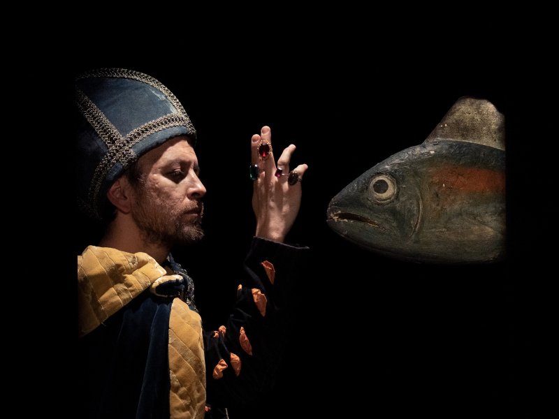 Mann in historischem Gewand mit hervorstechendem Hut, steht vor dunklem Hintergrund einen großen Fisch gegenüber und hebt seine mit Ringen besetze Hand in die Höhe.