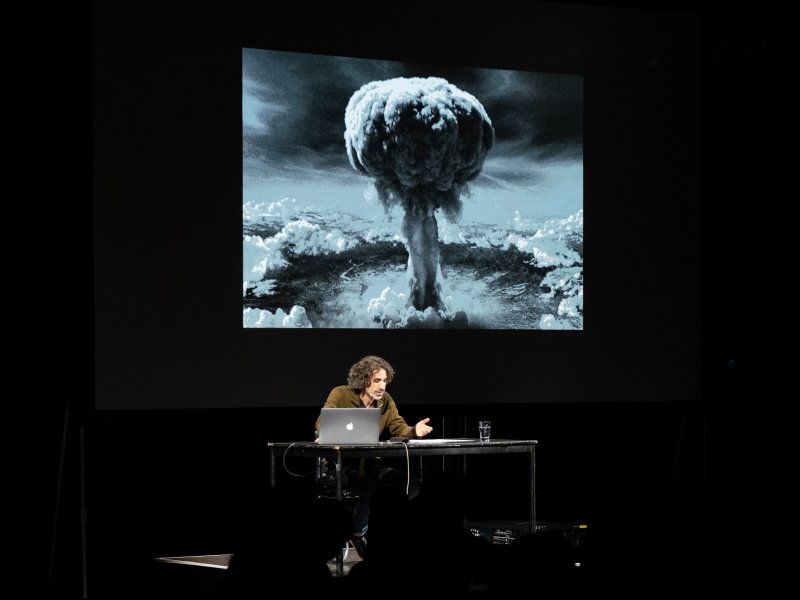 Ein Referent sitzt an einem Tisch mit Laptop, während auf die Wand hinter ihm das Foto eines Atomexplosionspilzes projiziert wird.