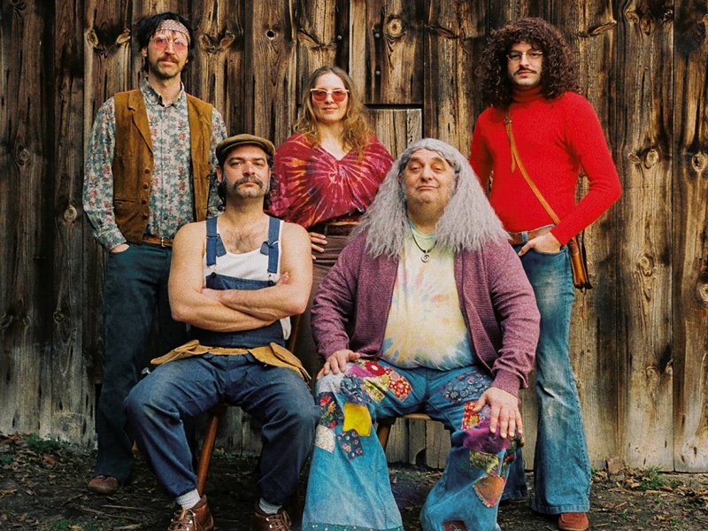 Fünf Personen in Hippie-Kleidung aus den 60er Jahren vor einer Holzwand. Zwei Personen sitzen im Vordergrund, drei stehen hinter ihnen. Sie tragen blaue Latzhose, Jeans, Batik-Shirts, bunte Hemden, Sonnebrillen und Lederschuhe.
