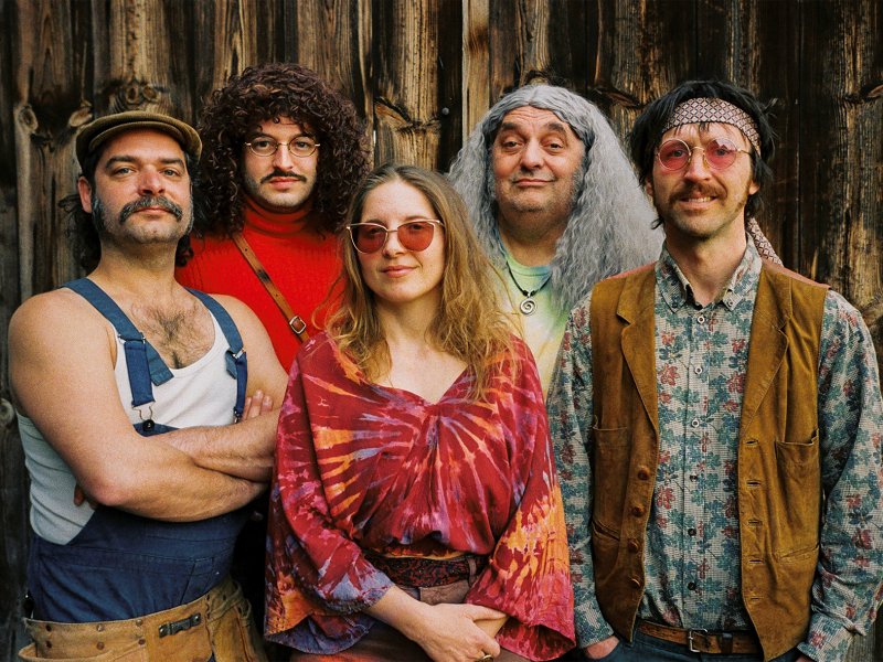 Fünf Personen in Hippie-Kleidung aus den 60er Jahren vor einer Holzwand. Sie posieren in einer Reihe und tragen blaue Latzhose, Jeans, Batik-Shirts, bunte Hemden, Sonnebrillen und Lederschuhe.