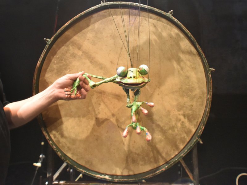 Eine Hand hält zwei Beine einer grüne Froschmarionette, die vor einer großen Trommel hängt. Die Marionette hat bewegliche Gliedmaßen und große Augen.