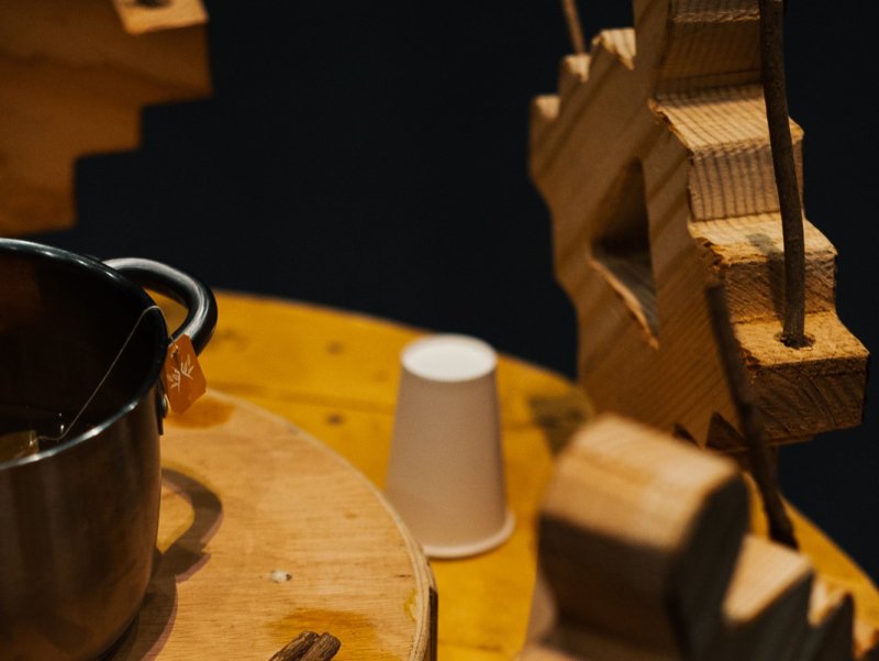 Holzfiguren um einen runden Tisch mit einem Topf voller Tee und einem weißen Becher. Die Figuren zeigen geometrische Männchen aus Holz.