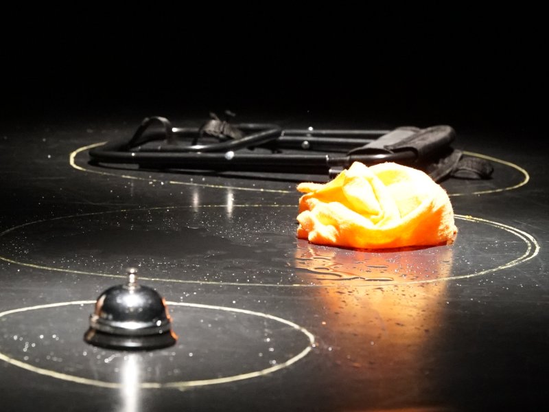 Ein orangefarbenes Tuch, eine Tischglocke und ein eingeklappter Stuhl liegen auf einem schwarzen Boden. Alle drei Gegenstände sind jeweils von einem hellen gezeichneten Kreis umgeben.