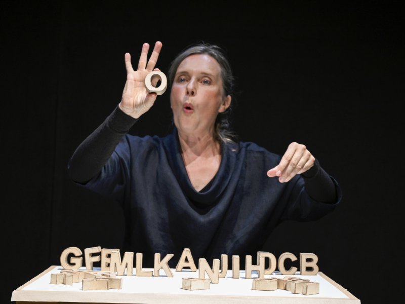 Frau in dunkler Kleidung, die einen aus Holz bestehenden O-Buchstaben hält, während sie eine Reihe von Holzbuchstaben auf einem Tisch zeigt. Mit ihren Lippen formt die einen O-Laut.