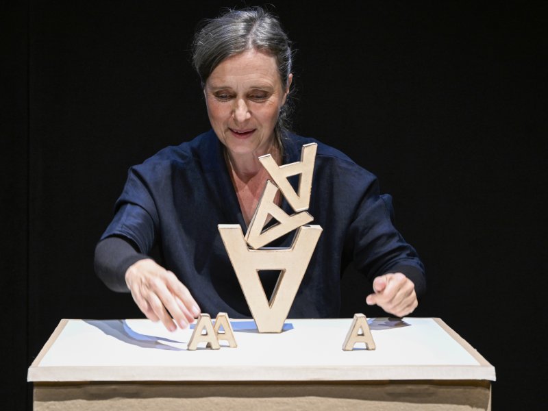 Frau in dunkler Kleidung und vor dunklem Hintergrund stapelt Holzbuchstaben auf einem Tisch.