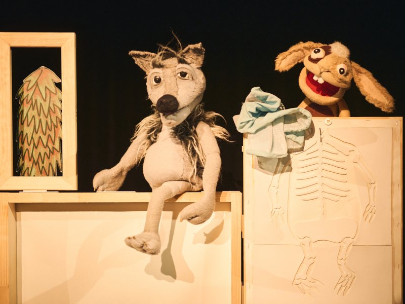 Puppenspiel mit zwei Figuren: ein grauer Wolf und ein brauner Hase. Der Wolf sitzt auf einer Kiste, der Hase steht hinter einer Holzplatte, auf der ein kleines Skelett eingeritzt ist.