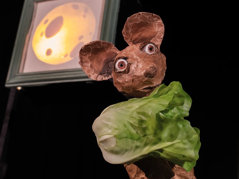 Pappmaché-Maus mit grünen Salat im Vordergrund vor einem Bild mit Mondmotiv im Hintergrund.