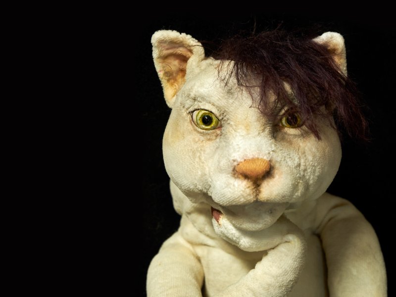 Eine plüschige weiße Katzenfigur, die eine Pfote an ihr Kinn legt. Die Figur hat gelbe Augen und braune Haare.