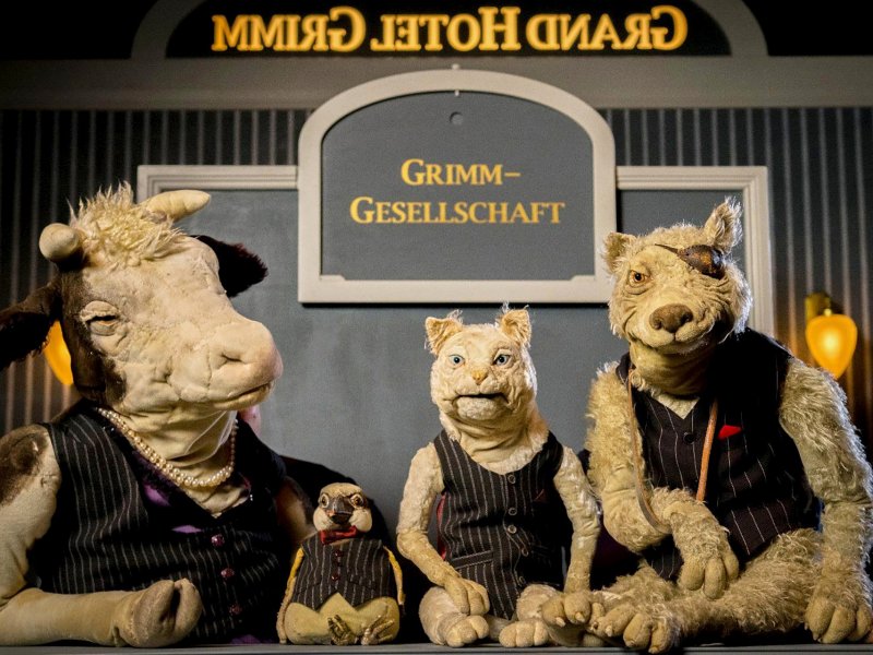 Tierpuppen in Nadelstreifenwesten, die verschiedene Tiere darstellen: eine Kuh, eine Katze, ein Vogel und ein Wolf. Sie posieren vor einem Schild mit der Aufschrift 'Grimm-Gesellschaft'.