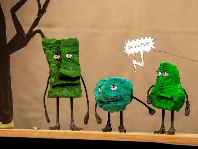 Drei bunte Figuren aus Papier und Draht stehen auf einem Tisch. Zwei Figuren in Grün und eine in Blau. Eine Figur spricht mit einer Sprechblase 'QUIIEEEK'.