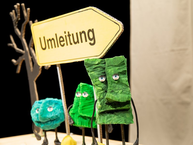 Drei bunte Figuren aus Papier in verschiedenen Grüntönen, stehen neben einem gelben Schild mit der Aufschrift 'Umleitung'. Im Hintergrund ein zweidimensionaler Baum aus Pappe.
