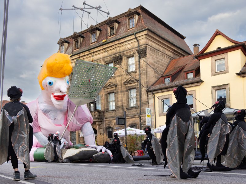 Menschen in Fliegenkostümen umgeben eine große aufblasbare Figur mit gelben Haaren und rotem Mund und einer Fliegenklatsche in der Hand auf einer Straße vor historischen Gebäuden.