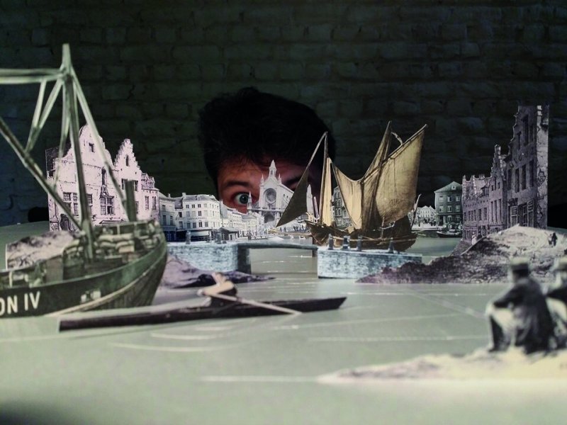 Kreative Kunstinstallation mit einer Miniaturlandschaft aus historischen Bildern, die aus Papier ausgeschnitten sind und die auf einem Tisch arrangiert sind, mit einer Person, die hinter der Szenerie hervorlugt.