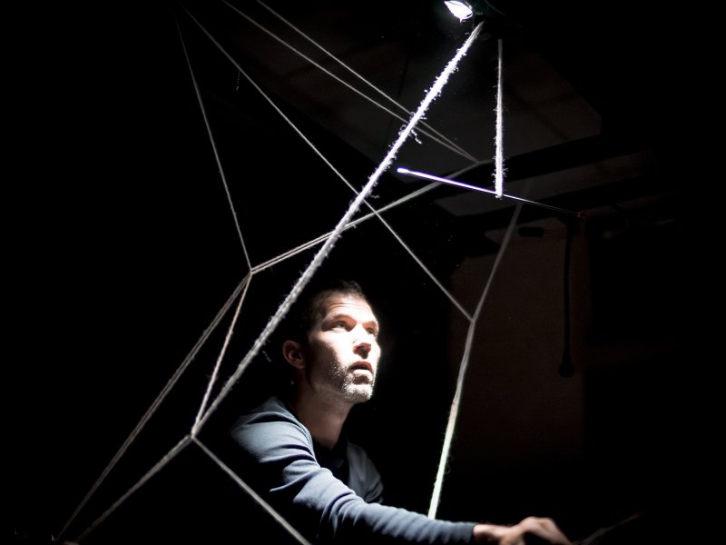 Mann in dunklem Raum, beleuchtet von einem Lichtstrahl, sitzt in einem geometrischen Netz aus Fäden und schaut nach oben.