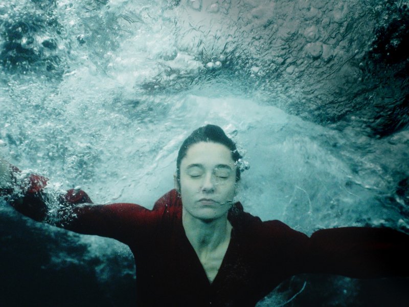 Eine Frau in rotem Kleid ist unter Wasser. Ihre Augen sind geschlossen und um sie herum sind viele Luftblasen.