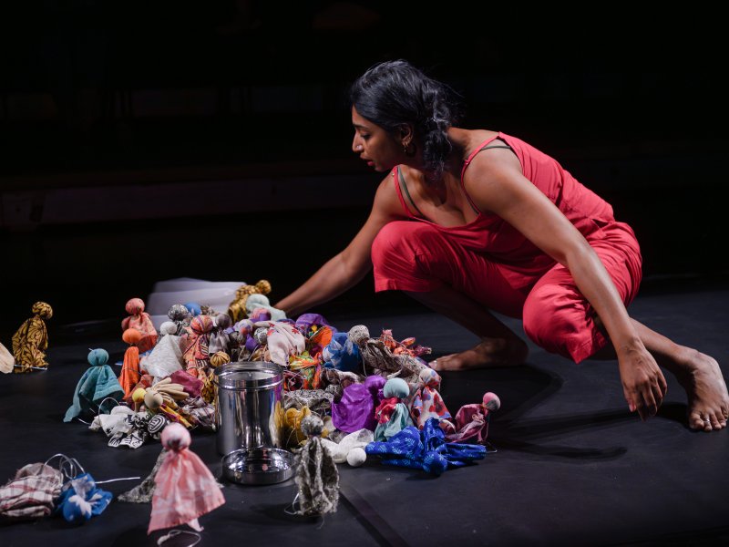Mallika Taneja sitzt in der rechten Bildhälfte in der Hocke auf einer schwarzen Bühne und vor schwarzem Hintergrund. Sie beugt sich zu unzähligen bunten Stoffpuppen herunter, die in einem Haufen zusammenliegen oder dahinter auf dem Boden stehen.