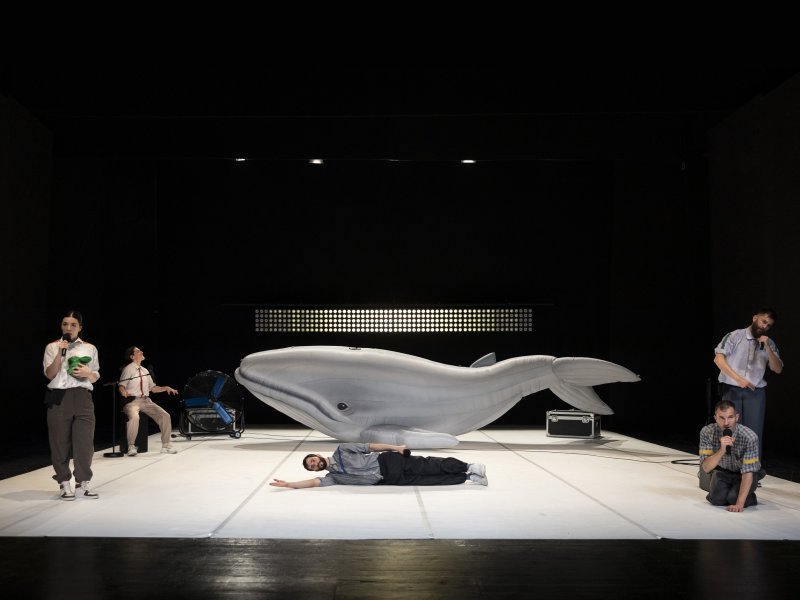 Theateraufführung mit einer großen Walskulptur und mehreren Schauspieler*innen in unterschiedlichen Posen, darunter ein liegender Schauspieler und halten oder sitzen an einem Mikrofon.