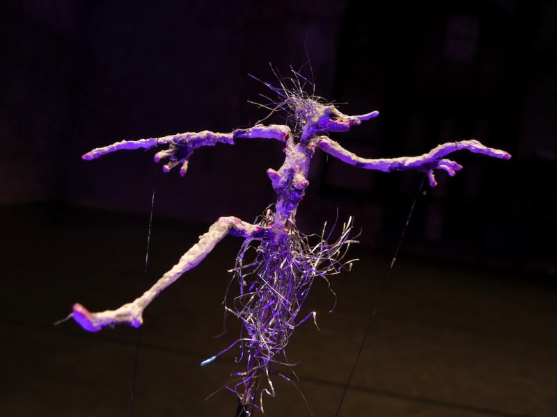 Skulptur eines stilisierten, humanoiden Geschöpfs mit langen Armen und spitzem Gesicht, aus organischen Materialien und Draht, im düsteren Hintergrund beleuchtet.