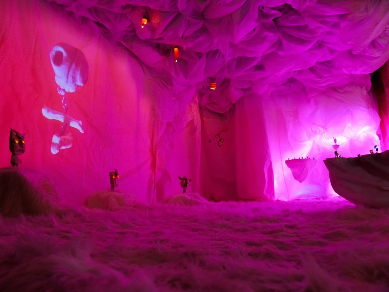 Futuristische Innenansicht eines Raumes mit rosa beleuchteten Wänden und einer Nebelartigen Bodenfläche, dekoriert mit schwebenden Lampen und Projektionen.