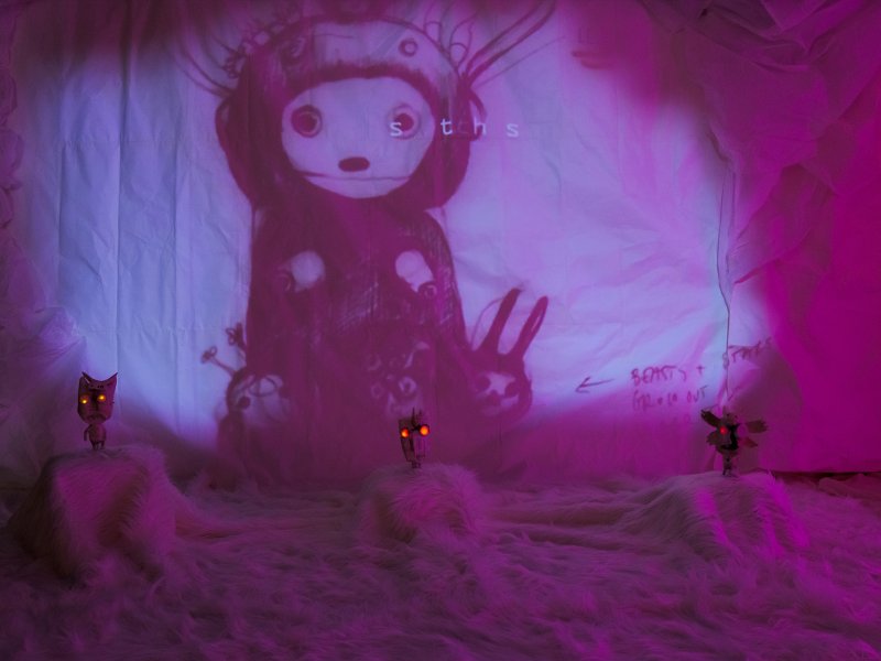 Bühnenbild mit einer Projektion eines fantasievollen, grafisch gestalteten Charakters auf einer weißen Leinwand, umgeben von kreativ gestalteten Objekten im Vordergrund, beleuchtet von rosa und lila Licht.