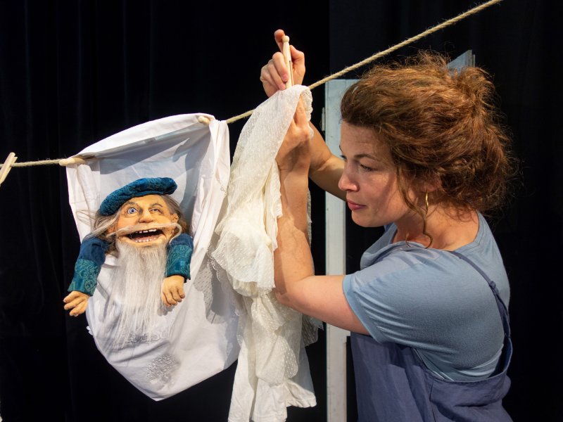 Frau mit braunen Haaren in einem blauen Oberteil, die eine Zwergenpuppe mit grauem Bart und blauer Mütze in einem Laken an einer Wäscheleine befestigt.