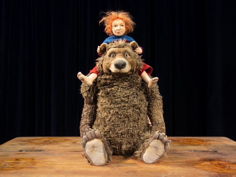 Ein zotteliger Bär mit großen Augen, der eine kleine, fröhliche Puppe mit roten Haaren auf dem Rücken trägt.