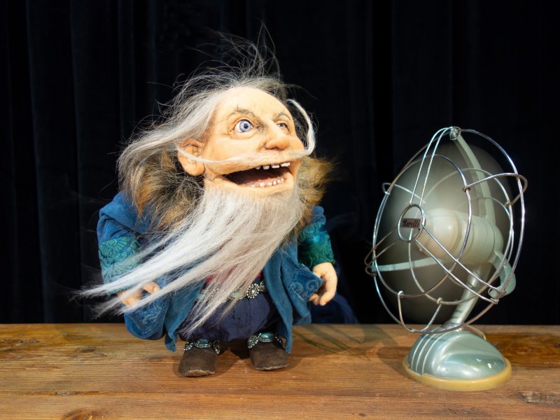 Puppe eines alten Mannes mit langem, grauem, wehendem Bart und beweglichem Gesicht. Sie steht neben einem Ventilator, der ihr Wind in ihr Gesicht bläst.