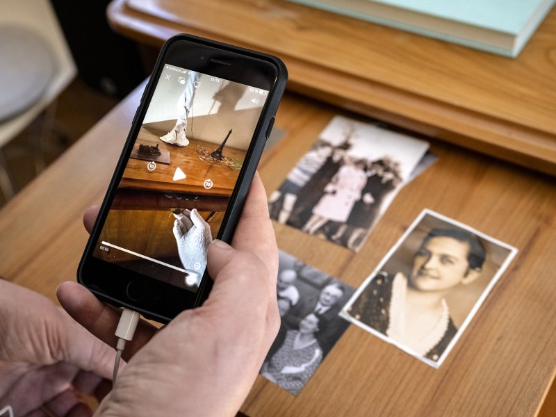 Eine Hand hält ein Smartphone, das ein Video zeigt. Auf einem Tisch liegen mehrere Schwarz-Weiß-Fotos.