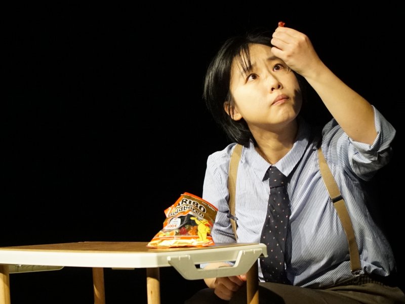 Schauspielerin in einem Hemd und Krawatte sitzt an einem Tisch, sieht nachdenklich auf ihre erhobene Hand, in der sie ein Gummibärchen hält. Auf dem Tisch liegt eine Packung Haribo-Gummibärchen.