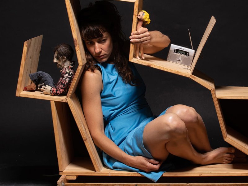 Eine Frau in einem hellblauen Sommerkleid, die vor einem dunklen Hintergrund in einer hausartigen Regalkonstruktion kauert. Mehrere Gegenstände wie ein Teekessel, ein Radio und zwei Puppen liegen auf den Regalbrettern.