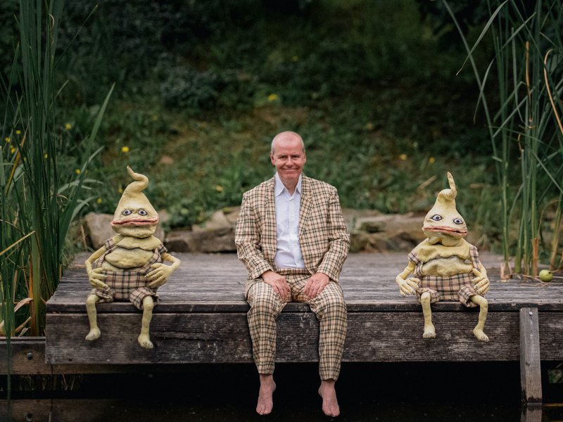 Ein Mann sitzt zwischen zwei froschähnliche Puppen auf einem Holzsteg. Im Hintergrund eine Böschung und Seegras.