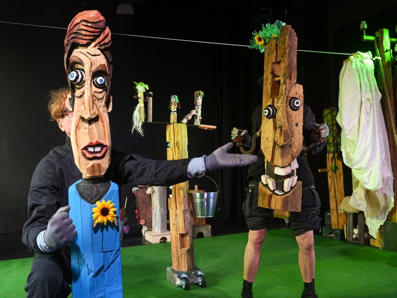 Zwei Personen mit Holzmasken auf einer Bühne mit grünem Fußboden. Im Hintergrund kleinere Holzfiguren und eine Wäscheleine.