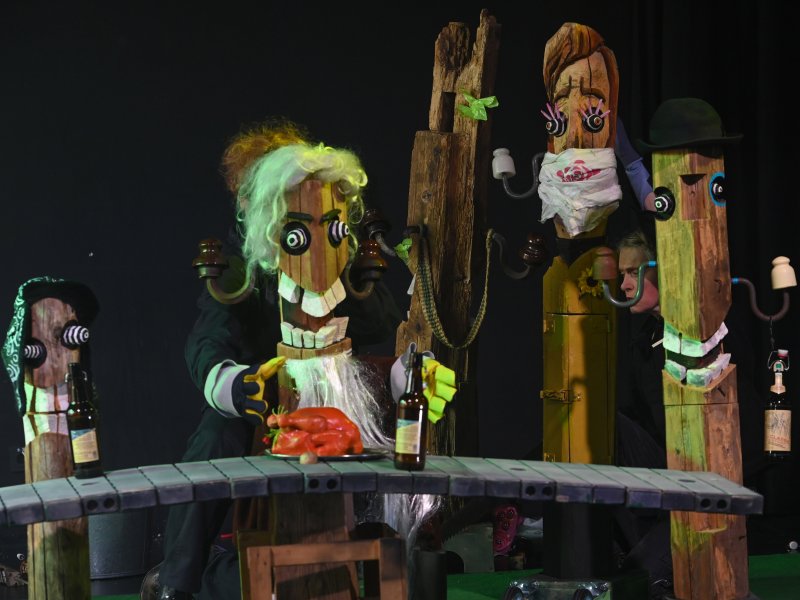 Vier Holzfiguren auf einer dunklen Bühne an einem Tresen, auf dem Bierflaschen und ein Braten stehen.