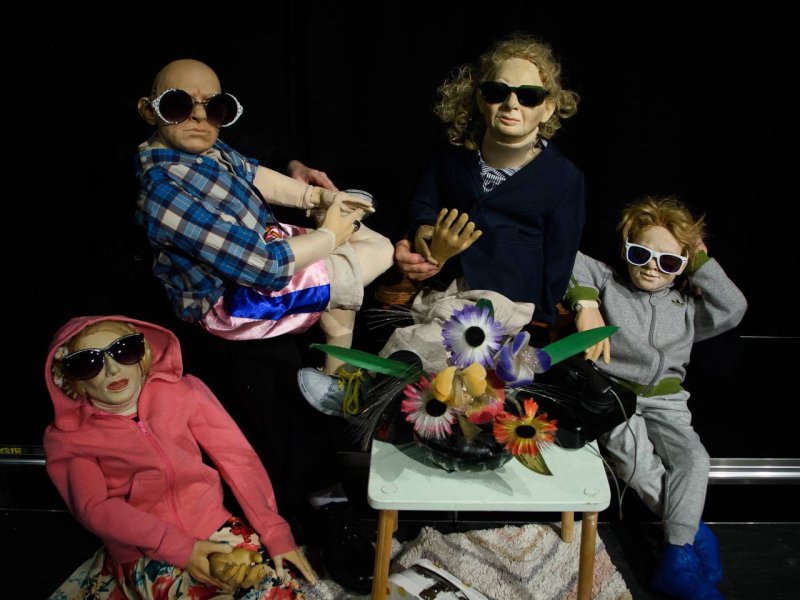 Fünf Puppen in Sonnenbrillen sitzen um einen Tisch mit einer Blumendekoration. Die Puppen haben unterschiedliche Kleidungsstile und Gesichtsausdrücke.