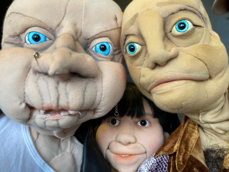 Drei Puppen mit großen blauen Augen und unterschiedlichen Gesichtsausdrücken, die nah beieinanderstehen.