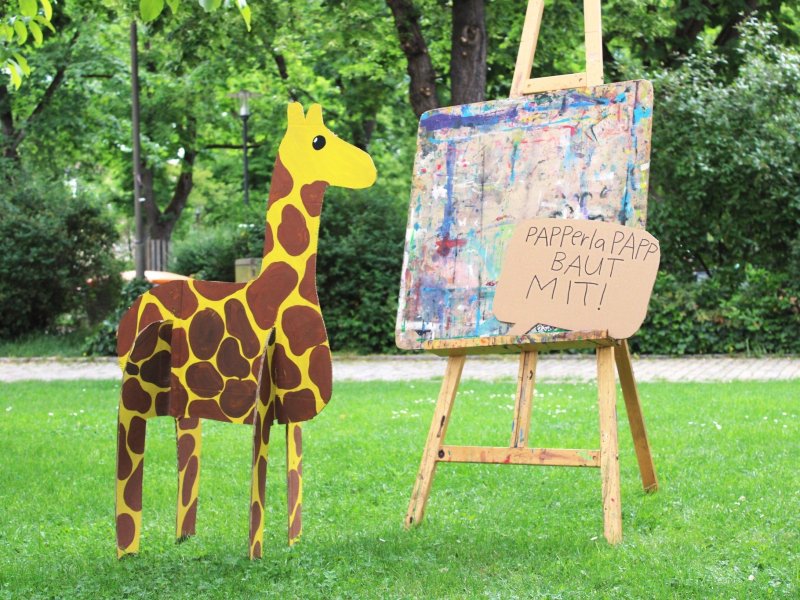 Eine Pappgiraffe steht neben einer Staffelei mit einem bunten Gemälde im Freien. Ein Schild fordert zum Mitmachen auf.