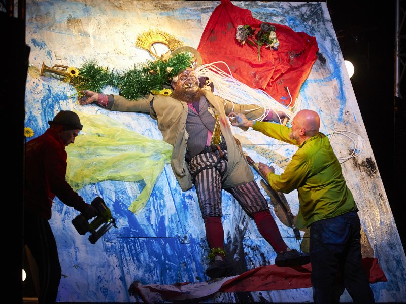 Theateraufführung mit einem Schauspieler, der an einer Wand befestigt ist, während zwei Personen mit Werkzeug und Farbdosen im Vordergrund arbeiten. Der Schauspieler trägt eine auffällige Kostümierung mit bunten Elementen und ist von dekorativen Pflanzen, Stoff, Seilen und einer Trompete umgeben.